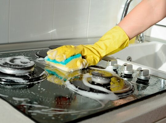 A hand in a yellow rubber glove washes a gas stove on a sunny day. Kitchen cleaning